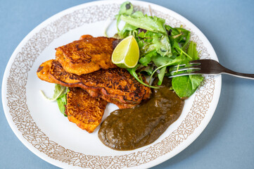 Chicken meat in coating and frying in batter, aubergine sauce and fresh green salad leaf with lime on plate.