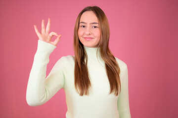 Portrait of a happy young girl showing okay gesture and winking isolated over pink background