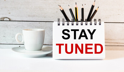 The word STAY TUNED is written in a white notepad near a white cup of coffee on a light background
