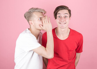 Close-up portrait of two young people, one whispering secrets to the other, shocked and very surprised, with their mouths wide open, isolated on a pink background. 