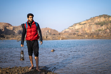 Young indian traveler standing near a lake in the mountains with a lantern, getting ready for camping in the night.