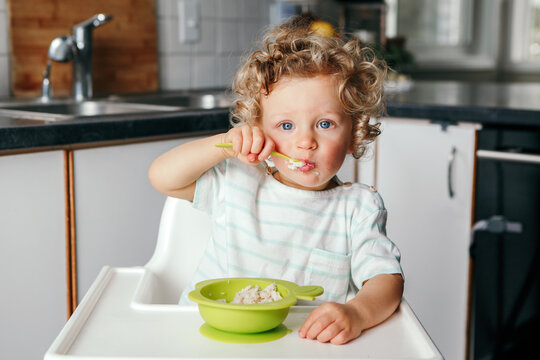 Cute Adorable Caucasian Curly Kid Boy Sitting In High Chair Eating Cereal Puree With Spoon. Healthy Eating For Kids Children. Toddler Eating Independently. Candid Real Authentic Moments.