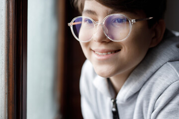 Portrait of young smiling boy in hoodie and glasses. Youngster expresses happiness. Teenager looking through the rainy window.