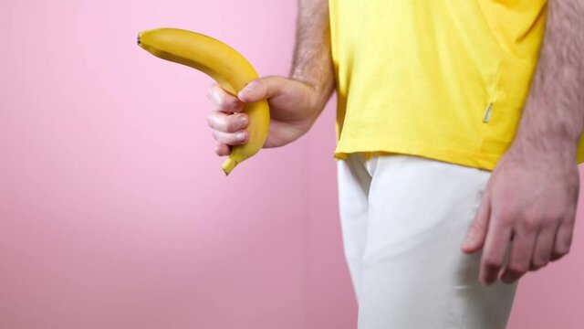 Potency. Man holds a banana in his hand at the level of the genitals, playing with it as with a gun. Close up. Pink background. The concept of men's health.