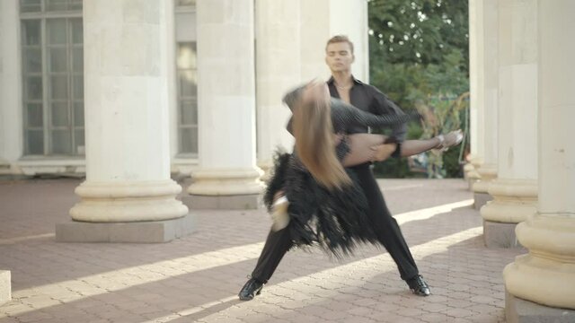 Wide shot of confident couple dancing paso doble between white columns in city. Professional man bending slim beautiful woman as column hiding dancers. Latin ballroom dance concept.