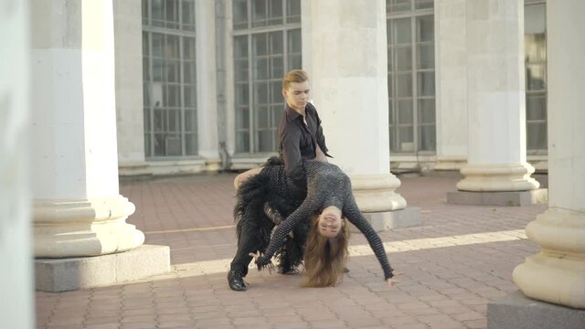 Wide shot of man catching and bending slim woman between urban columns. Portrait of professional Caucasian ballet dancers performing outdoors on sunny day.