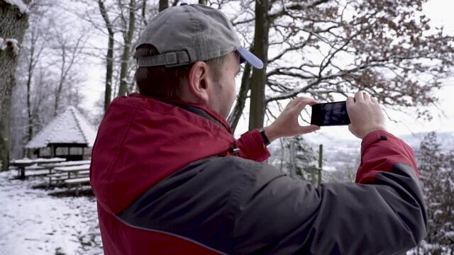 Static shot of a middle aged man in red winter jacket with gray cap in winter forest, filming beautiful view in Switzerland. Real time shot.