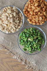 three glass bowls of multicolored roasted peanuts sit on a wooden table and a burlap tablecloth with