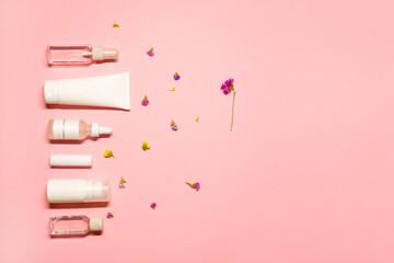 Flat lay setting with beauty goods: toner, lip balm, essential oil, deodorant, facial cream, and serum on a pink background with a copy space and colorful dried flowers.