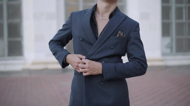 Stylish unrecognizable young Caucasian man buttoning up jacket outdoors. Confident rich businessman standing in urban city with building at background. Style and confidence concept.