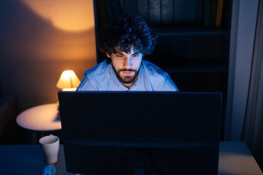 Close-up face of focused bearded man looking intently at monitor screen while working on computer at late night. Young caucasian freelancer businessman working and browsing internet in office.