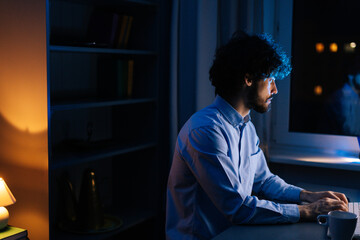 Side view of serious young business man working on computer in home at night. Focused freelancer looking device screen in dark living room at evening. Bearded male using pc at apartment nighttime