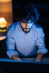 Top view of young man watching on monitor screen and working on computer at home office workplace at late night in dark room, tracking shot. Concept of remote working, distance learning.