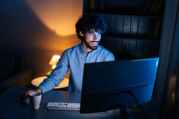Top view of happy smiling bearded young man looking intently at computer monitor screen while working on at late night. Concept of remote working, distance learning, Concept of remote working.