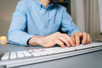 Close-up view of hands of unrecognizable man using computer and typing online message on wireless keyboard at light room sitting at desk. Closeup of working on desktop at late night. 