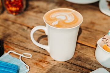 Closeup shot of white cup filled with aromatic coffee with heart shape latte art foam on  wood table near window with light shade on tabletop at cafe with face mask and glasses besides it, New normal