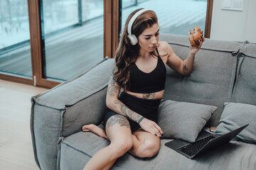 Obraz premium Beautiful sportswoman in black clothing with headphones and hamburger looks at laptop and sits on sofa in apartment.