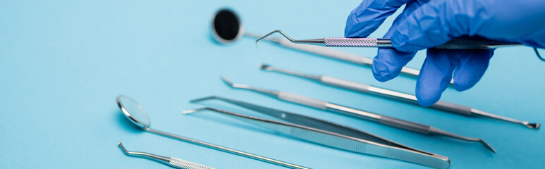 Cropped view of dentist in latex glove holding tool near dental set blurred on blue background,...