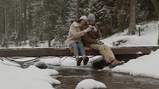 Wide shot of young African man and mixed-race woman sitting together on small bridge under stream in winter forest, drinking coffee from paper cups and enjoying view