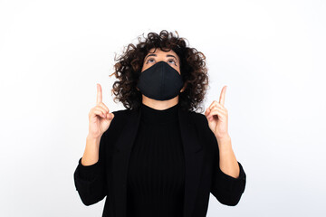 young beautiful caucasian woman wearing medical mask standing against white wall being amazed and surprised looking and pointing up with fingers showing something strange.