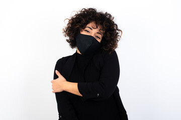 Fototapeta premium People, lifestyle, youth and happiness concept. Shy pretty young beautiful caucasian woman wearing medical mask standing against white wall,, feeling happy hugging herself.