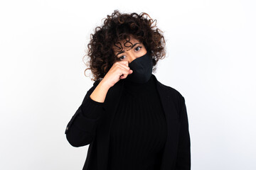 Unhappy young beautiful caucasian woman wearing medical mask standing against white wall, crying while posing at camera whipping tears with hand.