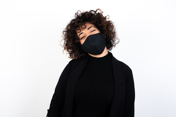 young beautiful caucasian woman wearing medical mask standing against white wall, with broad smile, shows white teeth, feeling confident rejoices having day off.