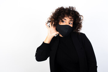 Oh my God! Funny astonished young beautiful caucasian woman wearing medical mask standing against white wall, opening mouth widely looking aside, with hand near ear trying to listen to gossips.