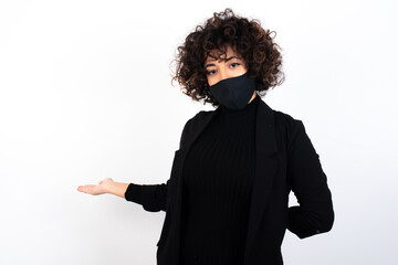 Portrait of young beautiful caucasian woman wearing medical mask standing against white wall,  with arm out in a welcoming gesture.