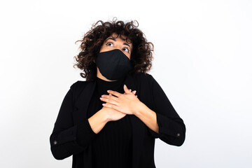 Happy smiling young beautiful caucasian woman wearing medical mask standing against white wall has hands on chest near heart. Human emotions, real feelings and facial expression concept.