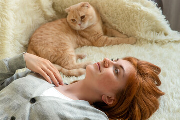 Pretty young romantic red-haired female lying on couch with domestic animal ginger cat and dreaming, looking up