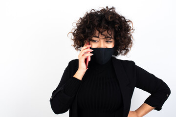 Portrait of successful joyful young beautiful caucasian woman wearing medical mask standing against white wall talking on mobile phone with friend. Lifestyle and communication concept