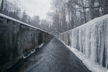 A stream in a concrete ditch or rainwater channel in winter in forest with long icicles on concrete walls. Storm drainage in winter forest with clear water.