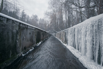 Fototapeta premium A stream in a concrete ditch or rainwater channel in winter in forest with long icicles on concrete walls. Storm drainage in winter forest with clear water.
