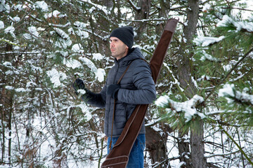 caucasian man in down jacket and jeans with shotgun in the case on the shoulder in the snowy forest