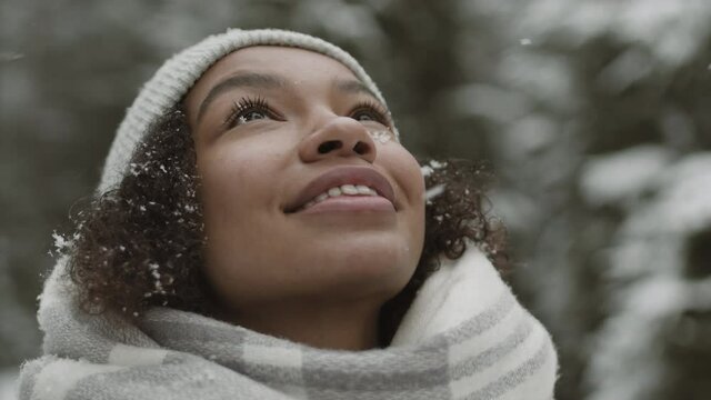 Close up of young mixed-race woman with curly hair standing outdoors, looking above and catching snowflakes with her face