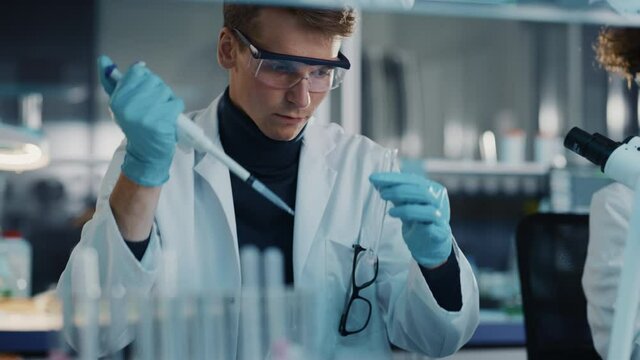 Modern Medical Laboratory: Young Caucasian Male Research Scientist Using Micro Pipette to Mix Chemical Liquids in a Test Tube. Microbiologists Conducting Biotechnology Research. Closeup Arc Portrait