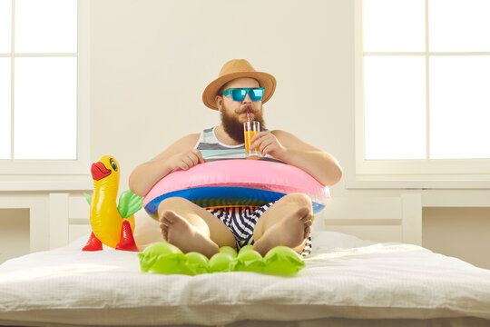 Funny young man in sunglasses and summer wear sipping beach cocktail, sitting barefoot in inflatable lifebuoy at home. Covid-19 quarantine, vacation in lockdown, canceled holiday travel plans concept