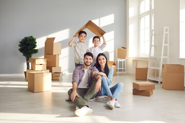 Fototapeta na wymiar Portrait of happy young family in new home. Smiling mom and dad sitting on floor with unpacked boxes, and children holding symbolic roof behind. Real estate, mortgage, buying and moving house concept