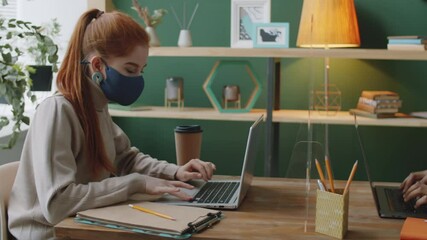 Side view of young Caucasian businesswoman and mixed race man in face masks sitting at office table with protective glass barrier and working on their laptops during covid-19 pandemic - Powered by Adobe