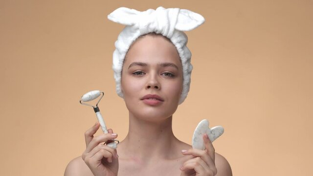 Beautiful young girl with beauty jade roller and guasha stone for massage on beige background. Cosmetic natural trend