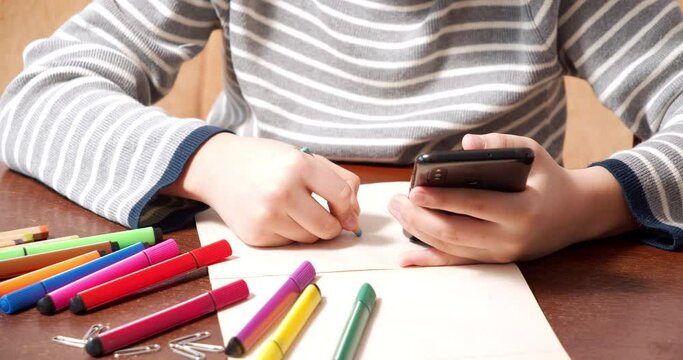 Close up of kid hands holding smartphone and drawing with colored pencil on paper. Online education and children's creativity concept