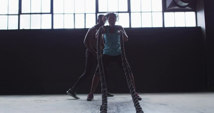 African american woman exercising battling ropes in empty urban building with man cheering her on