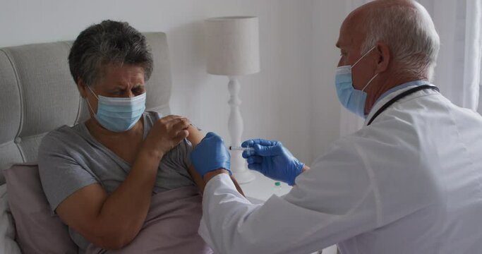 Senior caucasian male doctor vaccinating senior african american woman at home both wearing face mas