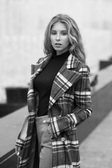 Fototapeta na wymiar Elegant woman walking city street. Fashion fall autumn look. Pretty caucasian blonde stylish girl wearing blue jeans, wine red pullover and squared wool coat. Female with makeup and wavy blonde hair