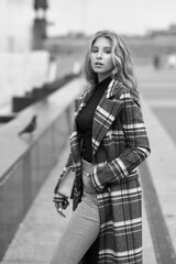Obraz premium Elegant woman walking city street. Fashion fall autumn look. Pretty caucasian blonde stylish girl wearing blue jeans, wine red pullover and squared wool coat. Female with makeup and wavy blonde hair