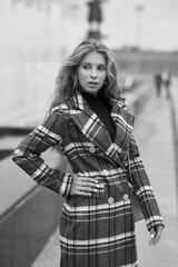Fototapeta premium Elegant woman walking city street. Fashion fall autumn look. Pretty caucasian blonde stylish girl wearing blue jeans, wine red pullover and squared wool coat. Female with makeup and wavy blonde hair