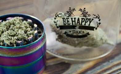 „Be happy all day” jar with marijuana buds and joint lie on a wooden background. Grinder near cannabis. Marijuana is medicine.