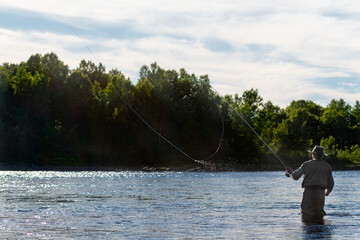 A male salmon angler stands in a river up to his waist with a long fishing rod wearing waders, fly...