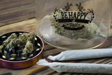 „Be happy all day” jar with marijuana buds and joint lie on a wooden background. Grinder near...